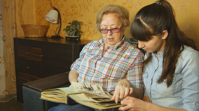 Old and young woman looking at family photo album on sofa at home