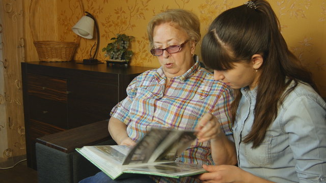 Old and young woman looking at family photo album on sofa at home