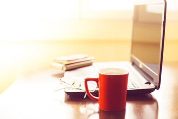 Coffee in a red cup on a table with a computer and notepads

