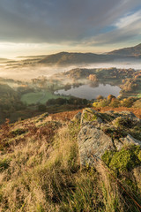 Lingering fog over Loughrigg Tarn on a fresh Autumn morning with mountain rocks in foreground.