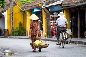 Unidentified old woman in traditional Vietnamese clothes carrying buskets with food on the street in Hoi An city. Hoian is recognized as a World Heritage Site by UNESCO. 
