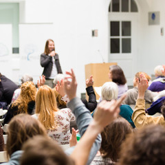I have a question. Group of people sitting at the chairs in conference hall, raising their hands....