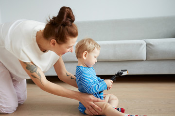 Young mother with her one years old little son dressed in pajamas are posing. Mom with son taking selfie on her smartphone or action camera in the bedroom.  Selective focus. Casual lifestyle photo.