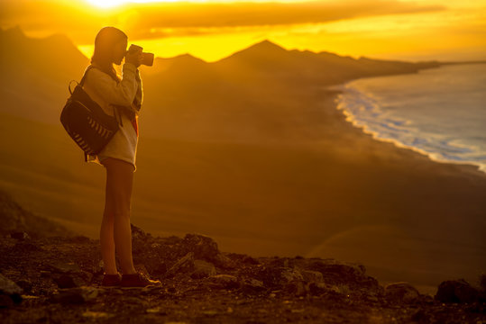 Young female traveler with backpack and photo camera photographing beautiful Cofete coastline with mountains on the sunset on Fuerteventura island in Spain. 