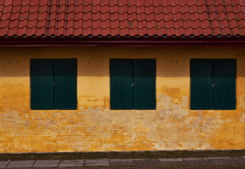 Three green wood shutters in a yellow plastered brick facade of an old house with red roof tiles