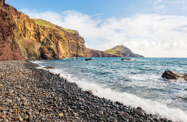 Volcanic black pebble beach with turquoise blue ocean water, high cliffs and blue sky during sunny day. Located on north coast of Ponta de Sao Lourenco, the eastern part of Madeira Island.