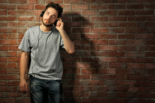 Young handsome man in grey T-shirt listening music with headphones on brick wall background