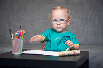 Умный мальчик в очках сидит за столом на котором стоит стакан с цветными карандашами 
