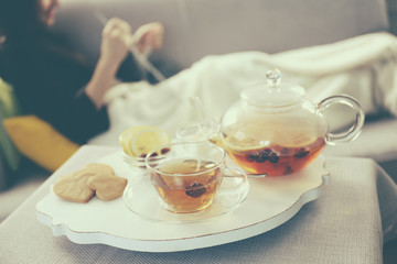 Tea cup and kettle on white wooden mat beside sofa on the knitting woman background