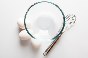 Glass transparent bowl with metal whisker and three white eggs aside directly from above