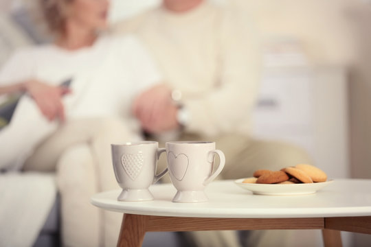 Two cups of tea with cookies on the table and mature couple on background
