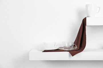 Set of cups and saucers with napkin on shelf against white wall background