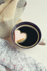 Cup of hot coffee with heart marshmallow and warm scarfs on light wooden background, close up