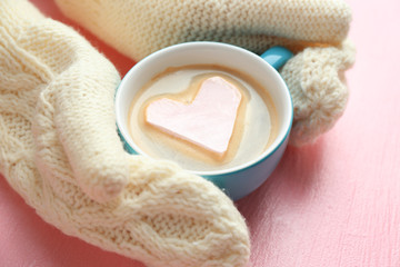 Female hands in warm mittens holding cup of hot cappuccino with heart marshmallow on pink background, close up
