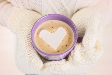 Female hands in warm mittens holding cup of hot cappuccino with heart marshmallow, close up