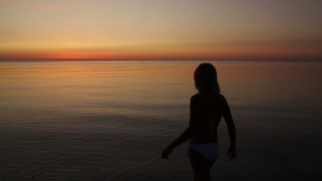 Young slim girl in white swimsuit enters the sea at sunrise with flying bird and dimples on the water (720p, 50fps)