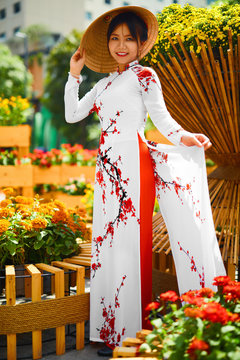 Culture Of Asia. Beautiful Happy Smiling Young Asian Woman Wearing Traditional White Ao Dai Dress ( Clothing ), Vietnamese Conical Hat ( Non La ) Leaf Hat In Flower Garden In Ho Chi Minh City, Vietnam