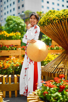 Traditional Clothing Of Vietnam, Asia. Beautiful Happy Asian Girl Dressed In National Traditional Ao Dai Dress ( Costume ), Vietnamese Conical Hat ( Non La, Leaf Hat ) In Flower Garden. Culture.