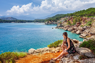 Naklejka premium Portret pięknej opalonej kobiety siedzącej na wybrzeżu morza. Gorący letni dzień i jasne, słoneczne światło. Panoramiczny widok na brzeg morza w pobliżu Kemer, Antalya, Turcja.
