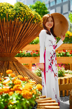 Culture Of Asia. Beautiful Happy Smiling Young Asian Woman Wearing Traditional White Ao Dai Dress ( Clothing ), Vietnamese Conical Hat ( Non La ) Leaf Hat In Flower Garden In Ho Chi Minh City, Vietnam