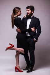 Portrait of young couple in love posing dressed in classic clothes on grey backround. Man with beard in Suit, Woman in black Dress and red shoes. The concept of Spring holiday