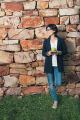 Fototapeta na wymiar Heatlhy smiling woman standing and leaning on rock wall, holding notepad and green apple, wearing blue glasses, casual jeans and sneakers, looking to one side.