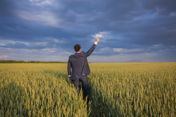 happy farmer, businessman, standing in wheat field over wind turbines background with his hands up and thumbs up