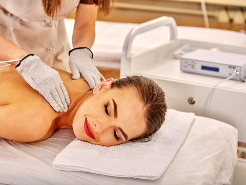 Young woman  with closed eyes lying on stomach receiving electric massage at beauty salon. Beautician wearing electricity gloves. In room is device for hardware cosmetology. 