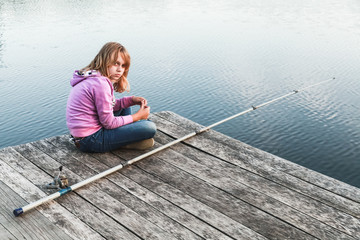 Fototapeta premium Blond girl sitting on a wooden pier with fishing rod