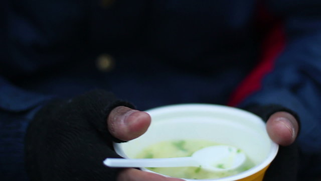 Miserable beggar warming cold hands with bowl of hot soup, charity kitchen meal
