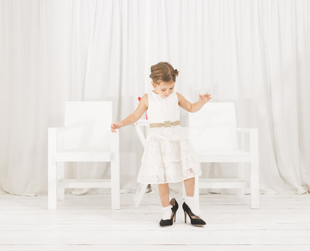 Adorable cute caucasian girl wearing white dress and black high heel shoes. Cute little girl trying to walk with big high heel shoes. Little girl fashionista in her mother's big heeled shoes