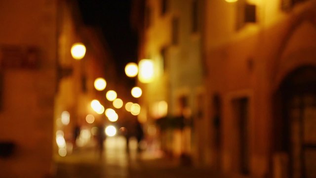  Pedestrians walking in medieval european Italy city street at  night with light illumination. Blurred defocused background.