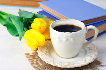 a Cup of coffee on the cloth with a little dish next to tulips and books on a white wooden table