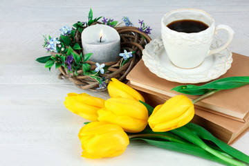 yellow tulips and a wreath with a candle inside lie next to books and a Cup of coffee on white wooden table