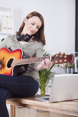 Pretty smiling brown haired girl sitting on desk playing some records on guitar while using laptop for support