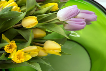Wiosenny bukiet kwiatów z żółtych i liliowych tulipanów oraz żonkili  na białym tle na zielonej szklanej misie paterze