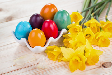 Pisanki, Wielkanocne dekoracje z kolorowymi jajami wielkanocnymi w ceramicznym stojaku, żonkilami na drewnianym sosnowym tle - obrazy, fototapety, plakaty