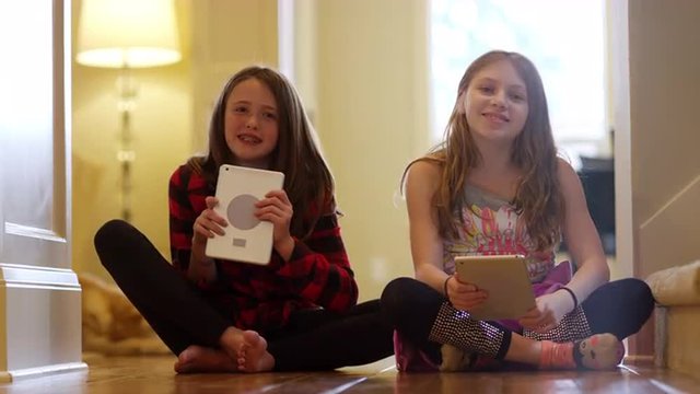 Two friends sitting together in the hallway smiling and then using their tablets