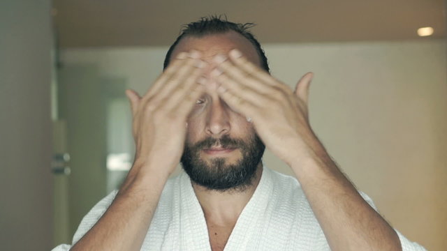 Young man in bathrobe applying cream on face in bathroom 
