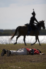 Napoleonic era soldier lies dead in a battle re-enactment whilst mounted soldier rides in background.