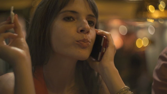 Young woman talking on cellphone, eating and drinking in cafe at night
