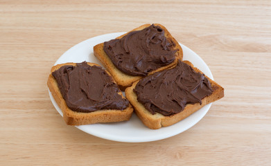 Chocolate peanut butter on hard toast on a white plate atop a wood table top.