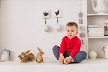 Little boy sits on the white kitchen buffet and plays with ceramic rabbits