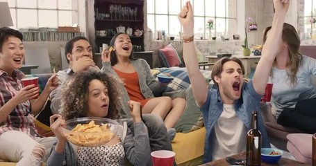 Diverse group of student friends arms raised celebrating goal watching sports event on TV together eating snacks  drinking beer  - Powered by Adobe
