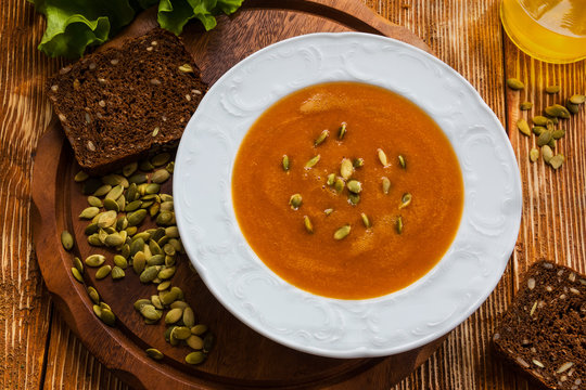 Pumpkin and carrot soup with seeds in a white plate on the wooden background. Green lettuce and olive oil.