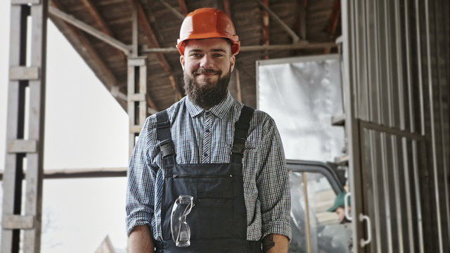 Portrait of cheerful bearded handsome young adult industrial engineer wear plastic helmet uniform against the background of loader. RAW video record.