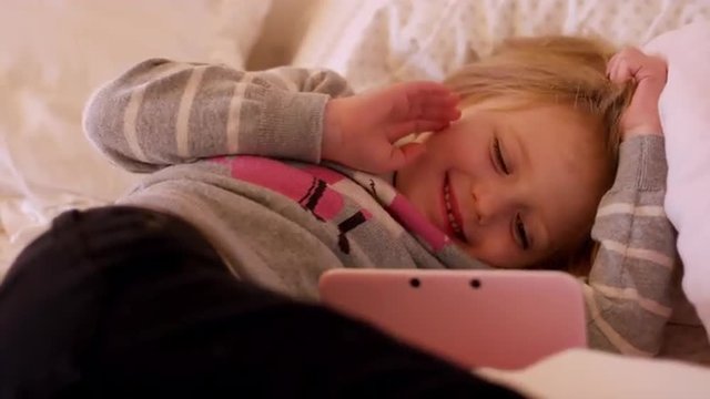A little girl laying on her bed watching a show on her handheld gaming system, and she smiles and waves