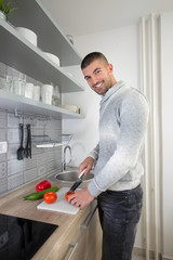 Handsome young man in the kitchen is preparing healthy meal from fresh organic vegetables. He is slicing tomato and looking at camera. Portrait composition