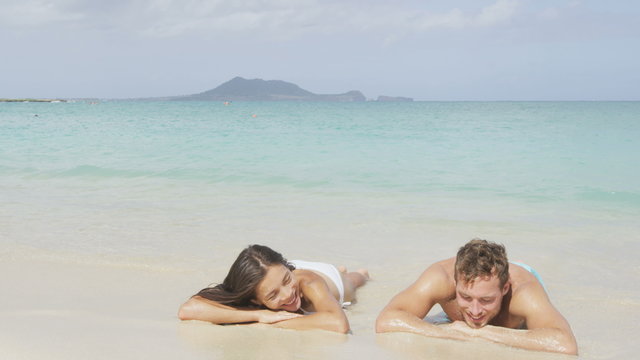 Beach vacations suntan couple relaxing in Lanikai, Oahu, Hawaii, USA. Blue sky and turquoise water on paradise beach  for holiday vacation travel concept. Multicultural woman and man.