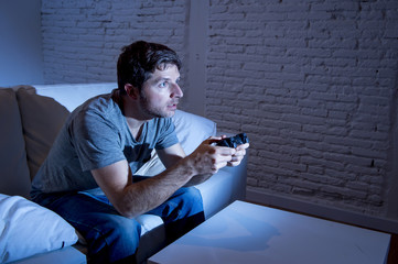 young excited man at home sitting on living room sofa playing video games using remote control joystick